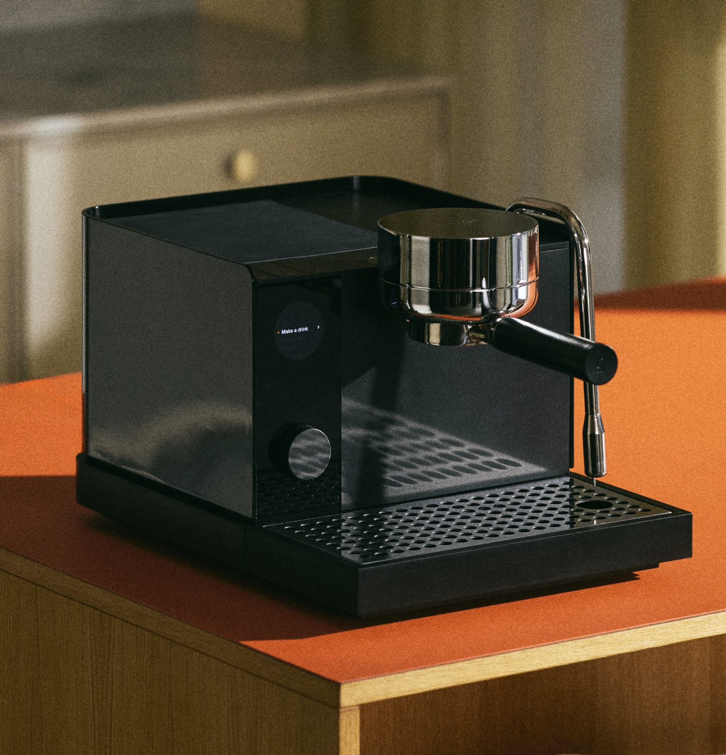 A sleek, modern black espresso machine with a metallic portafilter and steam wand sits on an orange countertop in a warmly lit kitchen.