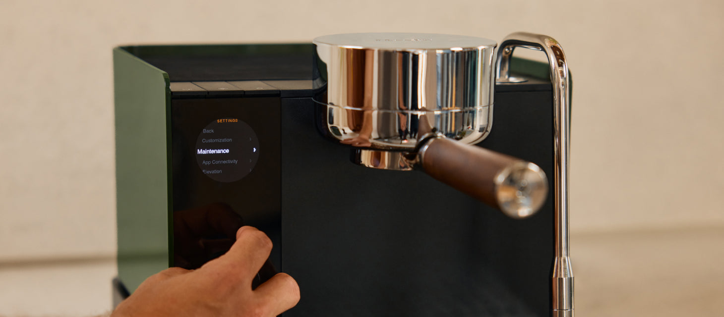 A person uses the touchscreen menu on a modern espresso machine with a chrome portafilter and steam wand. The display shows maintenance options.
