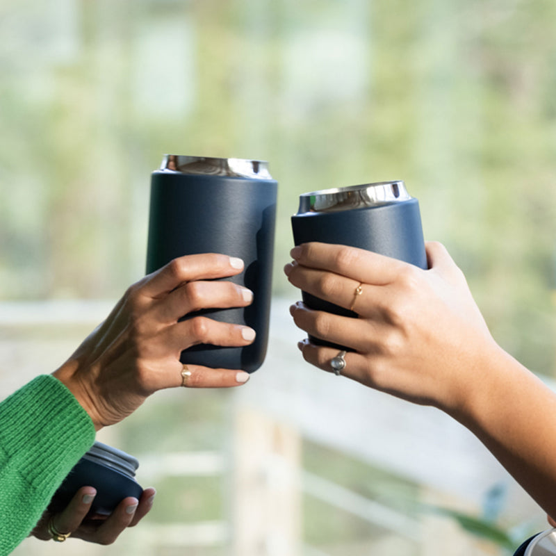 Two people toast with dark travel mugs, celebrating coffee moments amid blurred greenery.