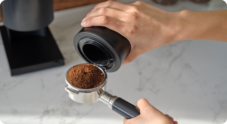 A home barista pours ground coffee from a container into a portafilter on a white counter.