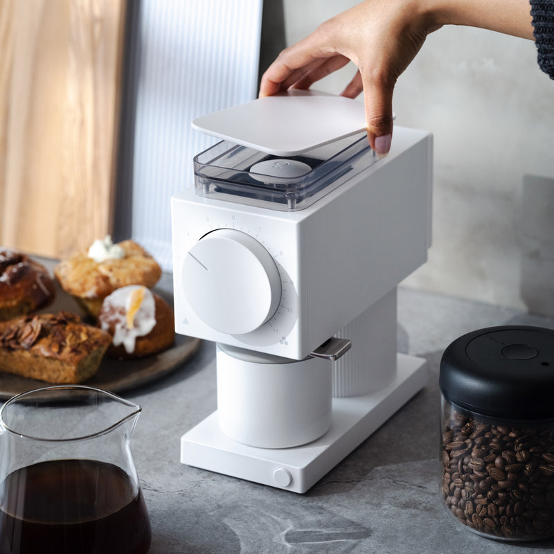 A home barista operates a modern white coffee grinder on a kitchen counter with pastries.