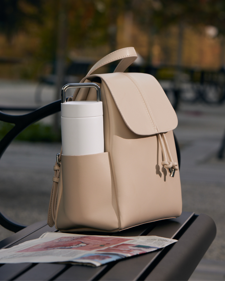 A beige backpack with a water bottle in its side pocket rests on a table, alongside a folded newspaper.